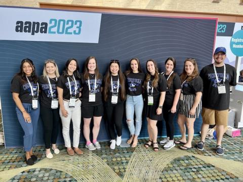 A photo of Andrew Allard, Nicole Clemente, Joelle Encarnacion, Melinda Hurteau, Giselle Lemus Tejada, Kadeja Miller, Jennifer Petrucci, Taylor Saimeri, Mark Sigwart, and Hailey Whipple (not in order), smiling in front of a dark-blue backdrop AAPA 2023 poster.